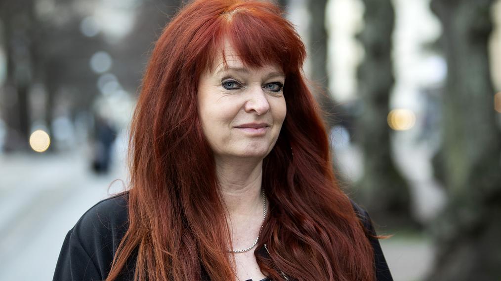 Lena Ulrike Rudeke står i stadsmiljö, hon har intensivt rött hår som det blåser lite i.