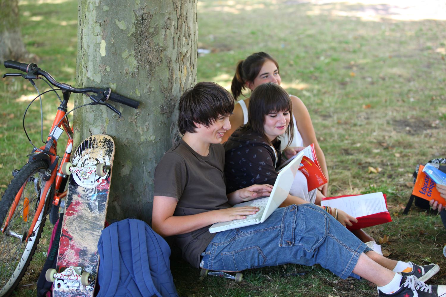 Glada tonåringar sitter på gräsmatta lutade mot en trädstam.