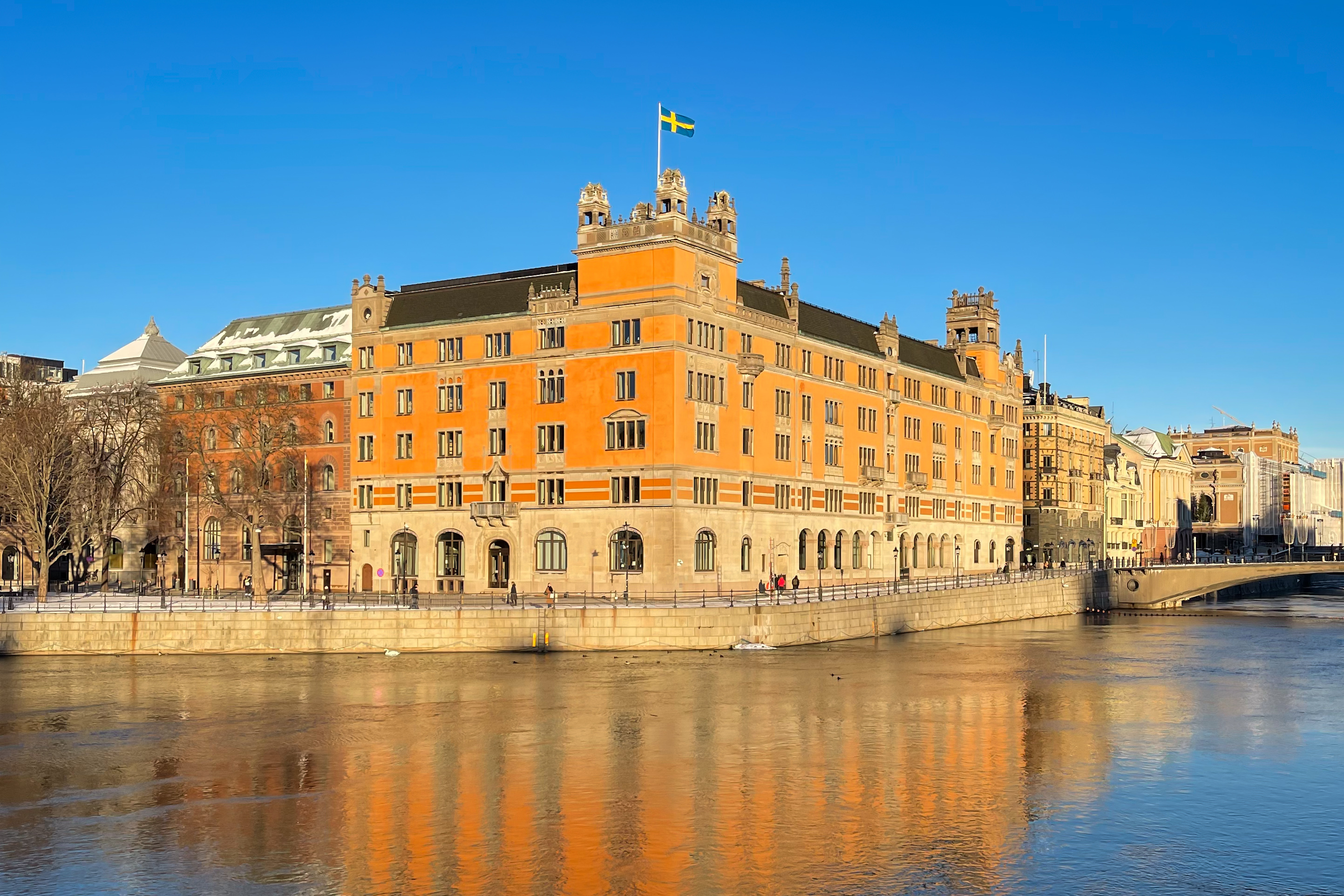 Rosenbad i vintrigt ljus. Svenska flaggan är hissad på taket och byggnaden speglas i vattnet framför.