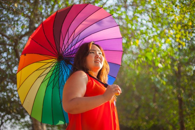 En glad kvinna under ett stort paraply i regnbågens alla färger.