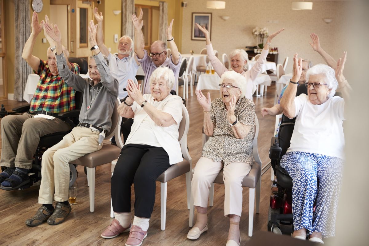 En grupp äldre i samlingslokal på äldreboende som applåderar och viftar med händerna. 
