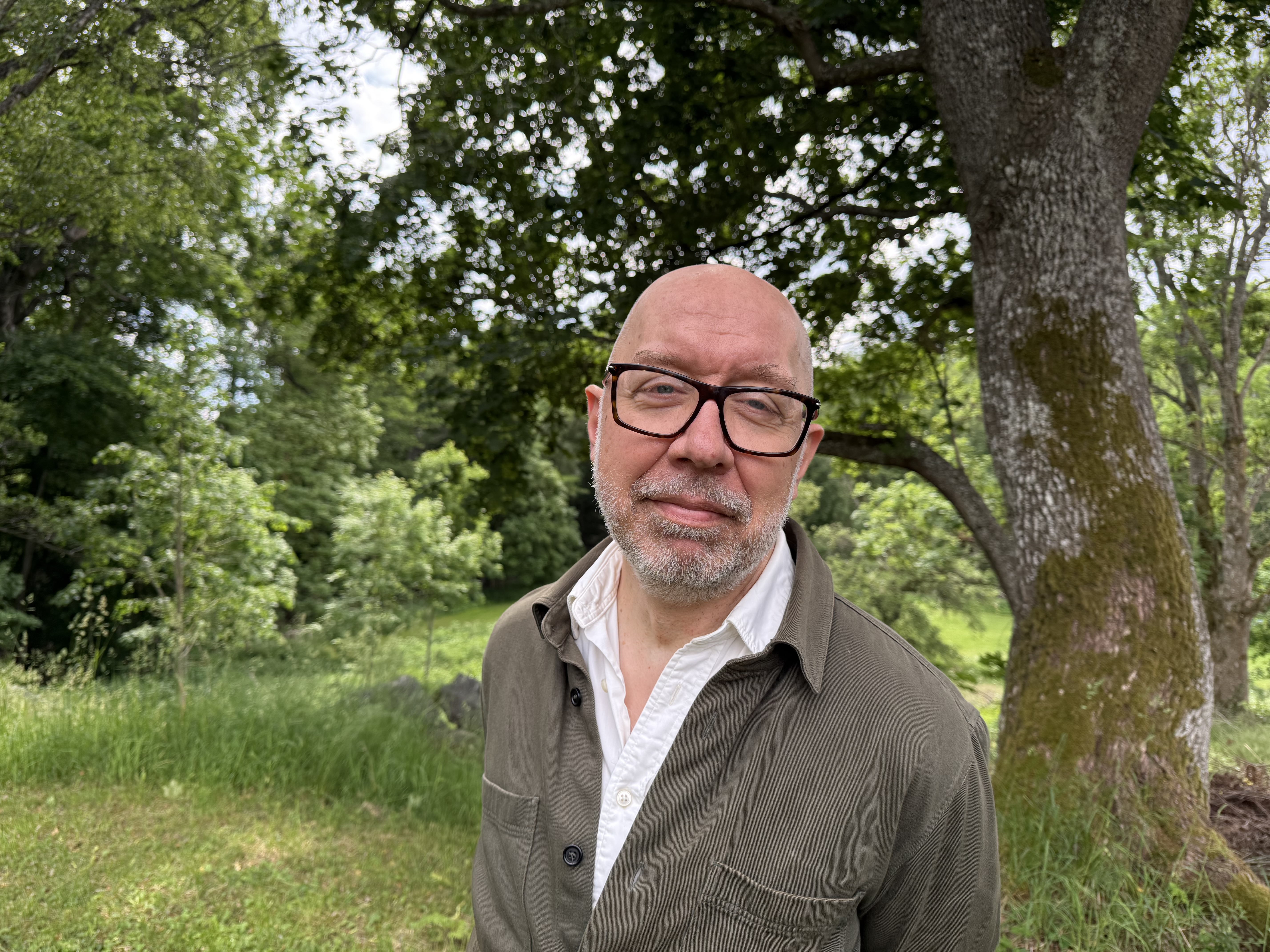 Ulf står med somrig grönska i bakgrunden, han har glasögon på sig och tittar in i kameran.