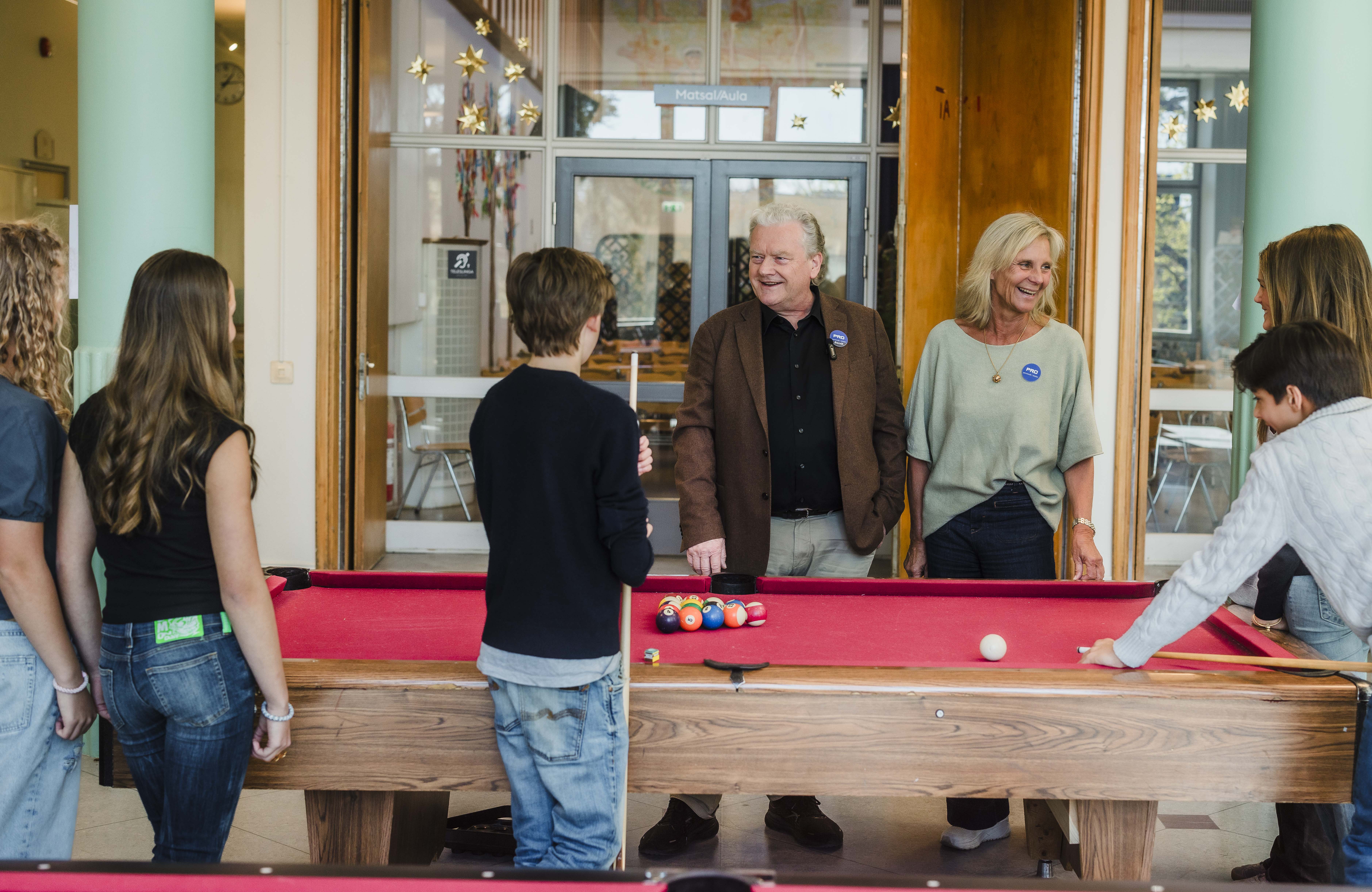 Två pensionärer står vid ett biljardbord med en grupp ungdomar. De ser glada ut.