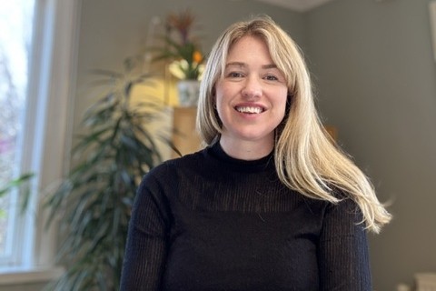 Felicia har långt blont hår som faller ut över hennes ena axel. Hon har en svart polotröja och ler stort rakt in i kameran.