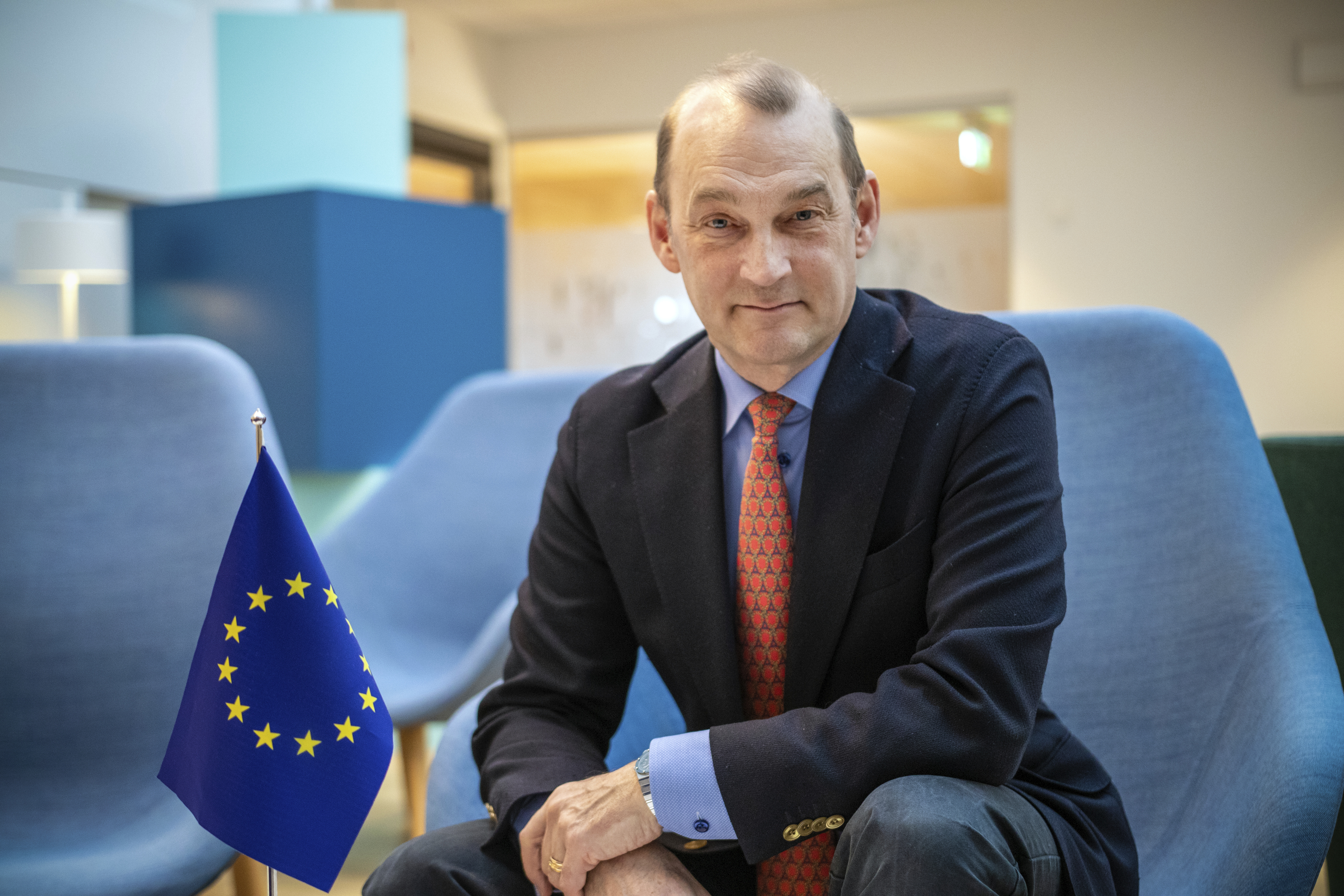 Mikael sitter i en blå fåtölj. Han tittar bestämt rakt in i kameran. I förgrunden syns en EU-flagga.