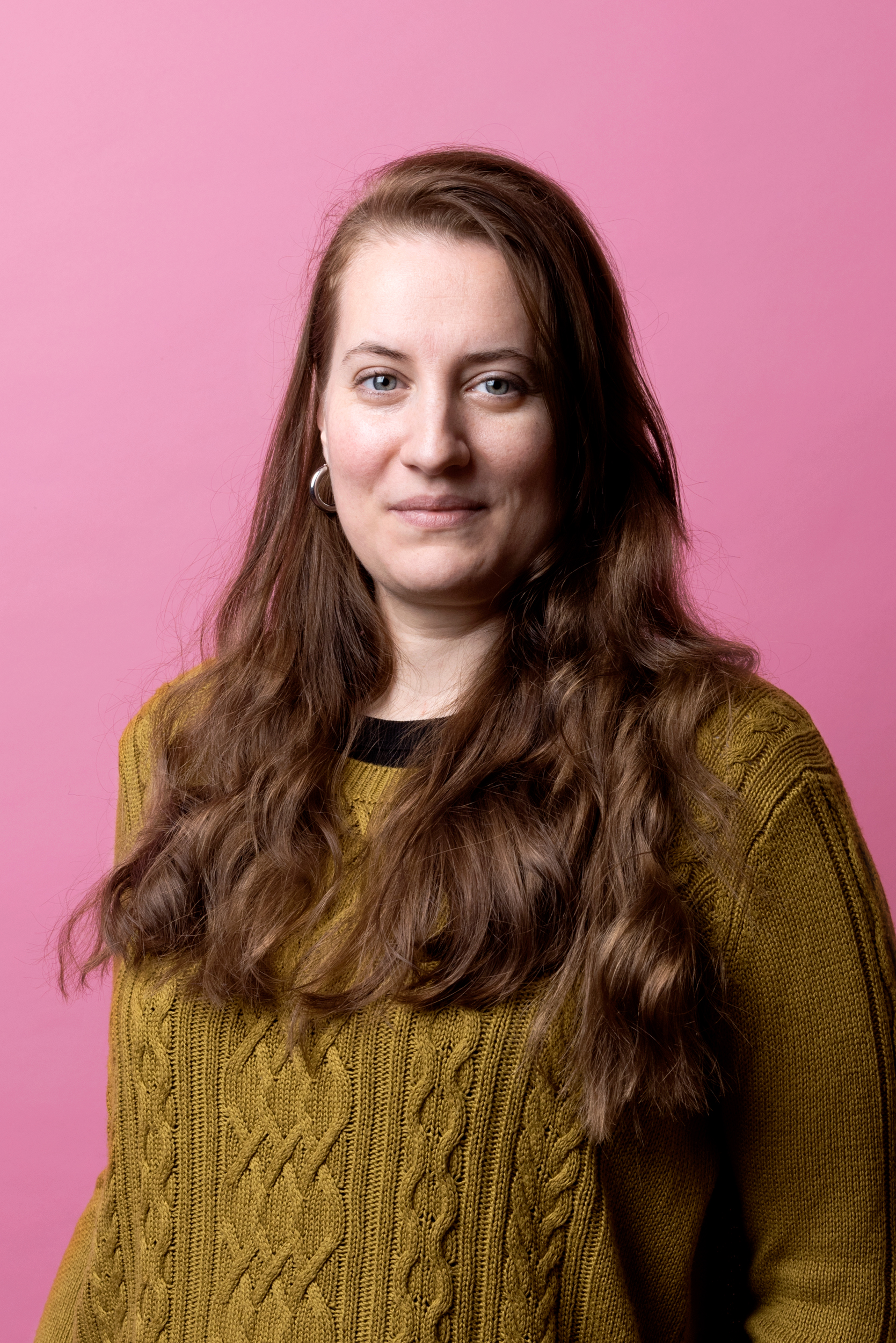 Karolina har långt brunt hår som faller ner över axlarna. Hon har en senapsgul stickad tröja på sig.