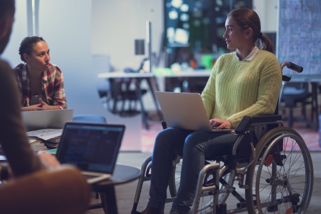 Ung kvinna i rullstol på möte, hon har en bärbar dator i knät.