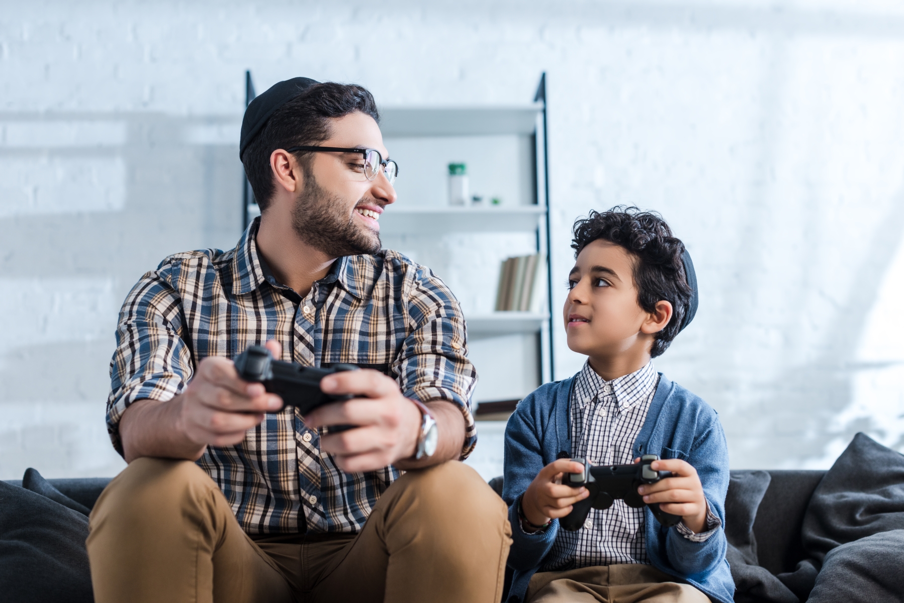 Judisk pappa och son sitter i en soffa, de ler mot varandra och spelar et tv-spel.