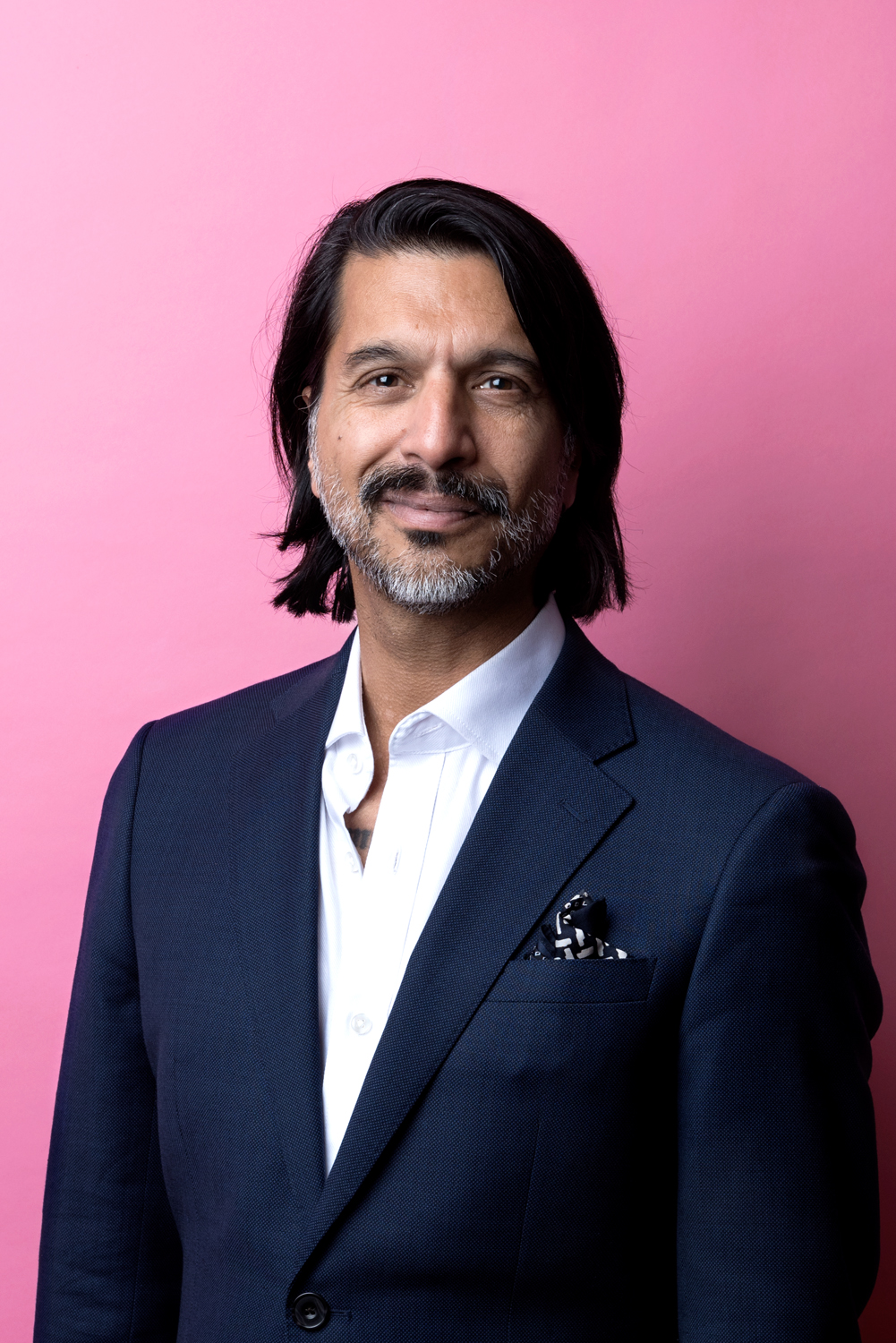 Porträttbild Qaisar Mahmood. Qaisar har axellångt mörkt hår, han är iklädd vit skjorta och mörkblå kavaj.